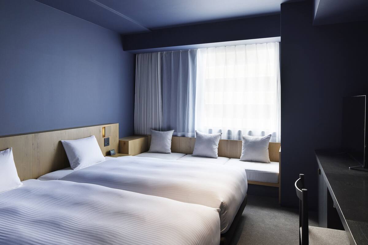 A hotel room with two beds, a large window with curtains, a wooden headboard, and a small bedside table with a lamp.