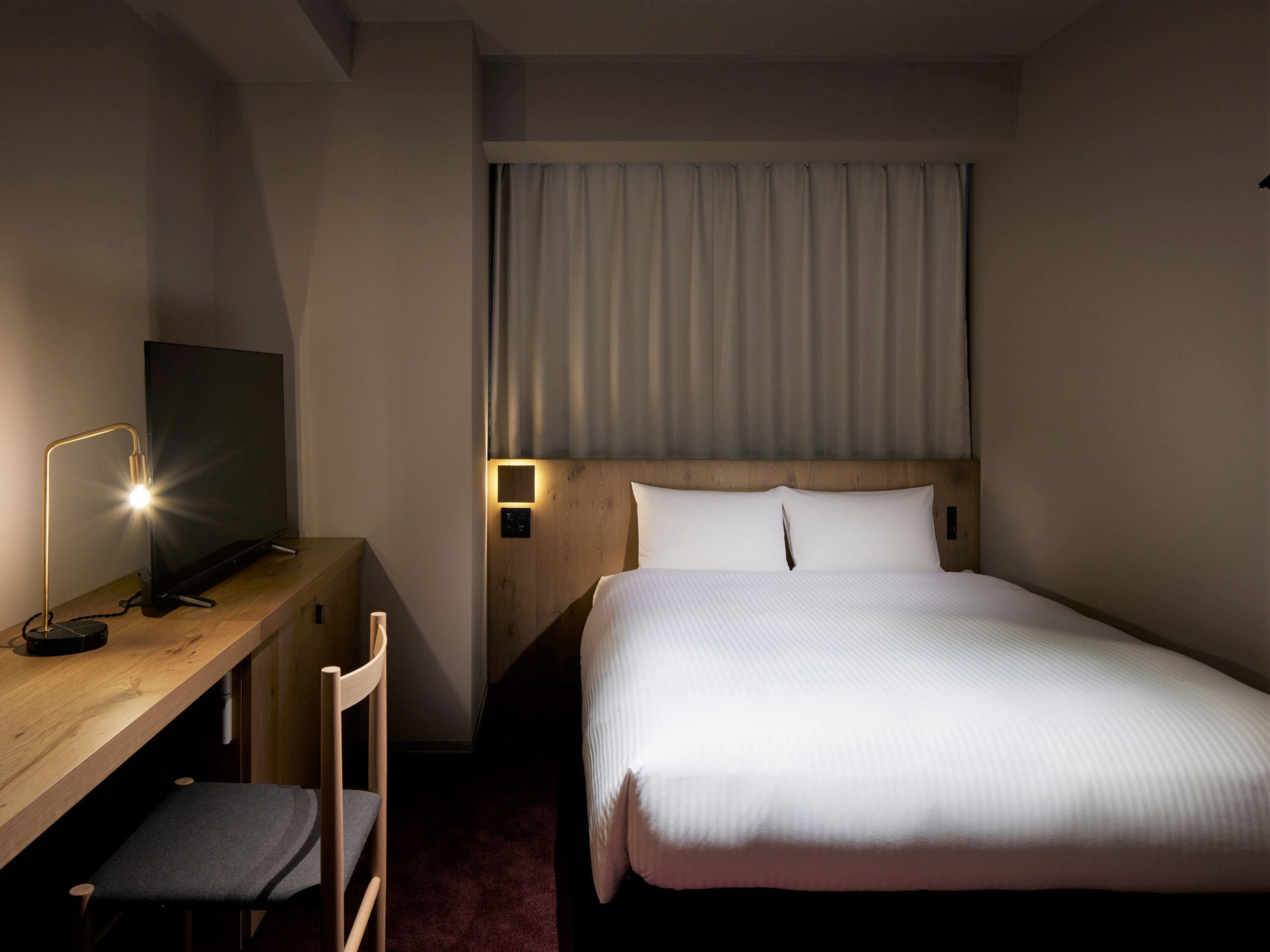 A hotel room with a bed, a desk with a chair, a television, and a nightstand with a lamp.