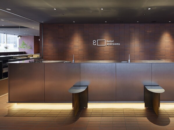 A modern hotel reception area with a reception desk, two seating areas, and a wall-mounted sign indicating hotel and rooms.