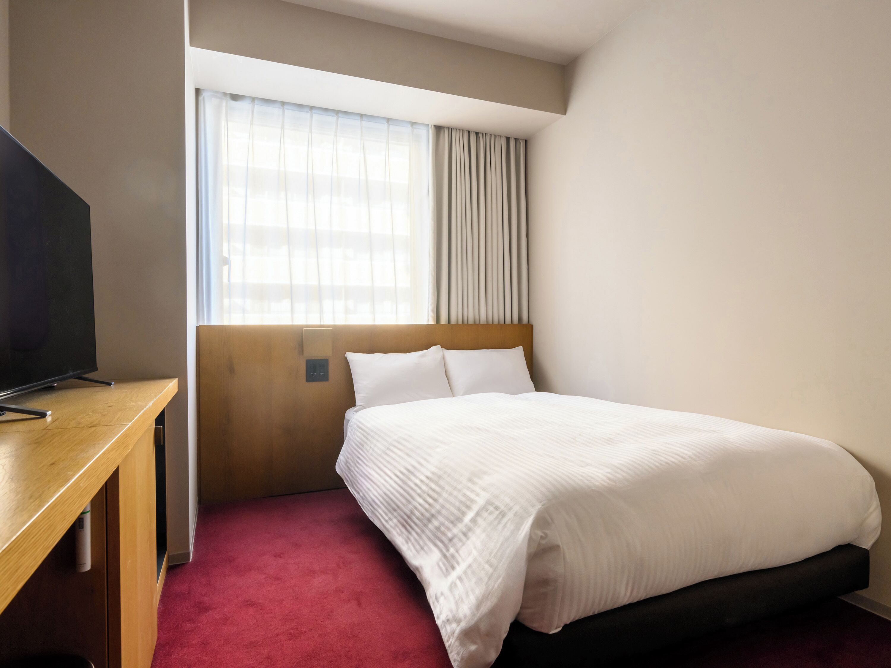 A hotel room with a bed, a TV on a wooden desk, and a window with curtains.