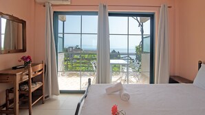 A bedroom with a large window, a wooden desk, a bed with a rolled towel, and a view of a balcony with outdoor furniture. - Apartment 'Andromaches Large' with Sea View & Shared Pool (Corfu)