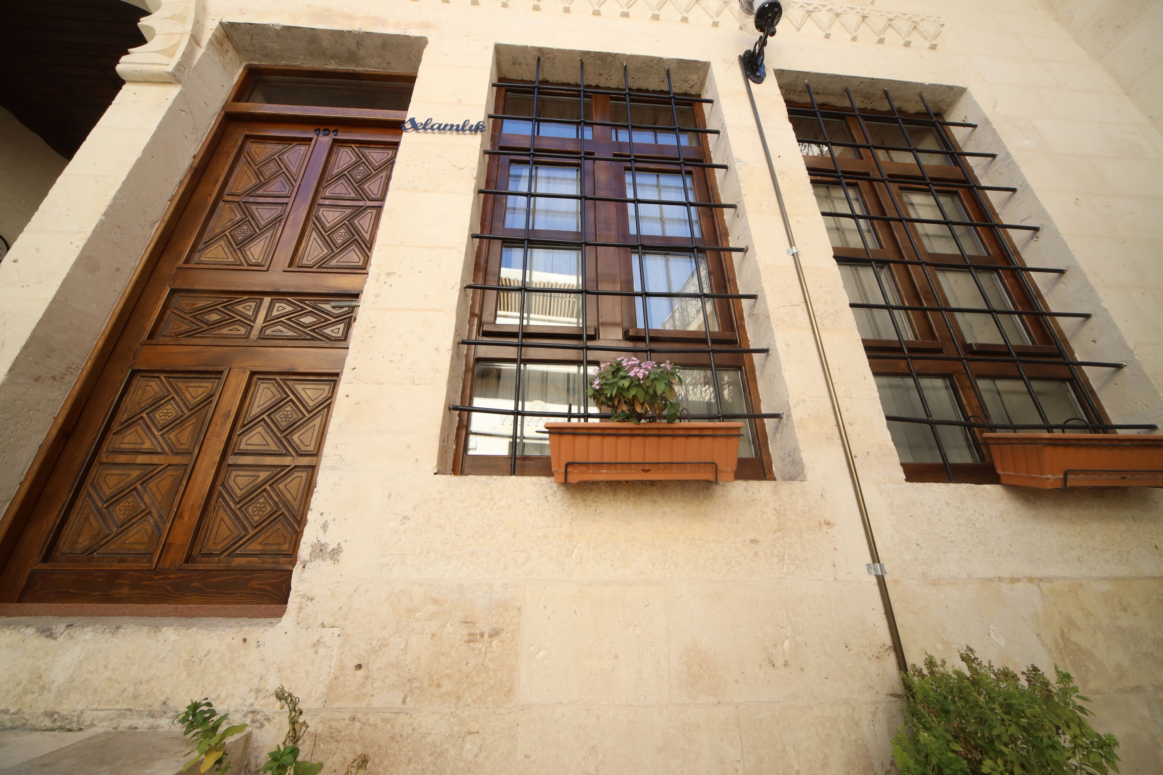 Fachada de edificio con grandes puertas y ventanas de madera, una maceta pequeña con flores y un cartel que dice 'Selamlik'.