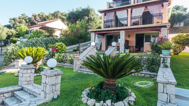A two-story house with a balcony, stone pillars, and a garden with plants and flowers.