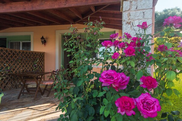 Une terrasse avec une table et des chaises en bois, une clôture en treillis et un rosier aux fleurs roses.