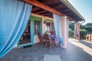 Um alpendre coberto com mesa e cadeiras, uma porta que dá para o interior e vista para o verde ao fundo.