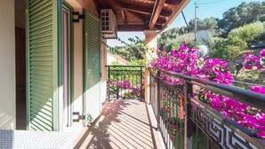 A wooden balcony with a metal railing, a green door, and a view of a garden with purple flowers. - 'Villa Pepy Luxury Apartment' with Mountain View, Garden & Wi-Fi (Corfu)