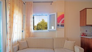 A room with a sofa, a window with a view of trees, a kitchen area, and a wall with a painting. - 'Villa Pepy Luxury Apartment' with Mountain View, Garden & Wi-Fi (Corfu)