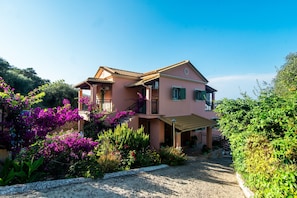 Une maison rose à deux étages, dotée d’un balcon, entourée d’une végétation luxuriante et de fleurs violettes.