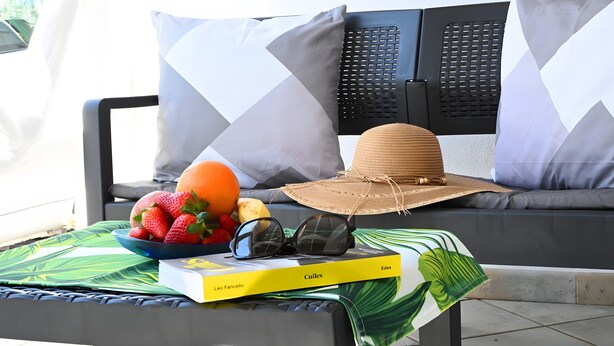 Un banc en métal noir, recouvert d’une nappe à motifs verts et blancs, un chapeau de paille, un bol de fruits, un livre intitulé « Edeos » de Leo Fancello, et deux coussins à motifs géométriques.