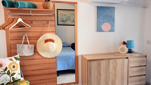 Une chambre à coucher avec un lit, une armoire en bois, un miroir, un chapeau accroché, une serviette et une commode avec une lampe.
