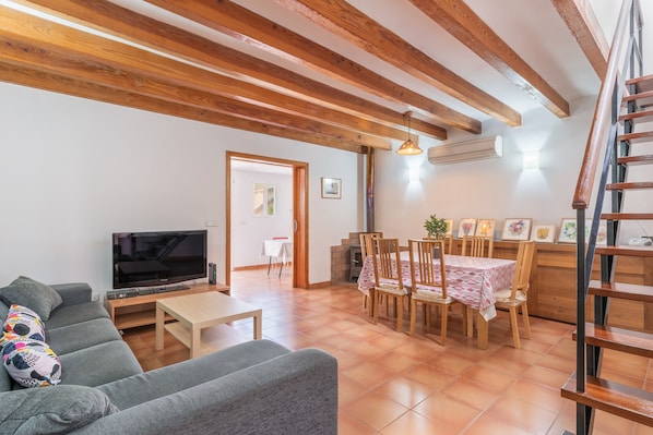 A living room with a sofa, TV, wooden beams, dining table, chairs, and a staircase.