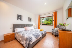 Camera da letto con un letto, comodino, cassettiera, finestra con tende e vista su spazi verdi.