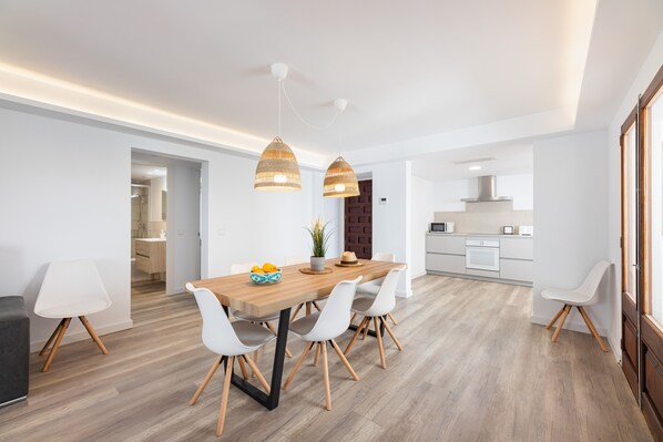 A modern dining area with a wooden table, white chairs, pendant lights, and a kitchen in the background.