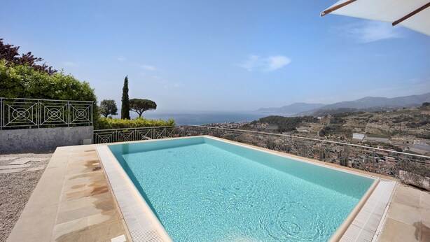 A rectangular outdoor swimming pool with clear blue water, surrounded by a stone deck and metal railings, overlooking a coastal landscape with hills and a view of the sea.