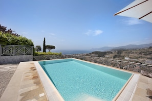 Una piscina rectangular al aire libre con agua azul cristalina, rodeada por una terraza de piedra y barandillas de metal, con vistas a un paisaje costero con colinas y una vista del mar.
