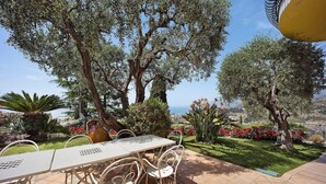 An outdoor patio with a table and chairs, surrounded by olive trees and a view of the sea.