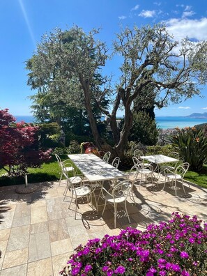 An outdoor dining area with a table and chairs, surrounded by trees and flowers, overlooking a coastal landscape.
