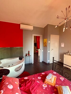 A hotel room with a large bed, a bathtub, a dining area with a red tablecloth, and a hallway leading to another room.