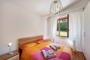 Un dormitorio con cabecera de madera, cama con ropa de cama naranja y rosa, una lámpara de noche y una ventana con cortinas.