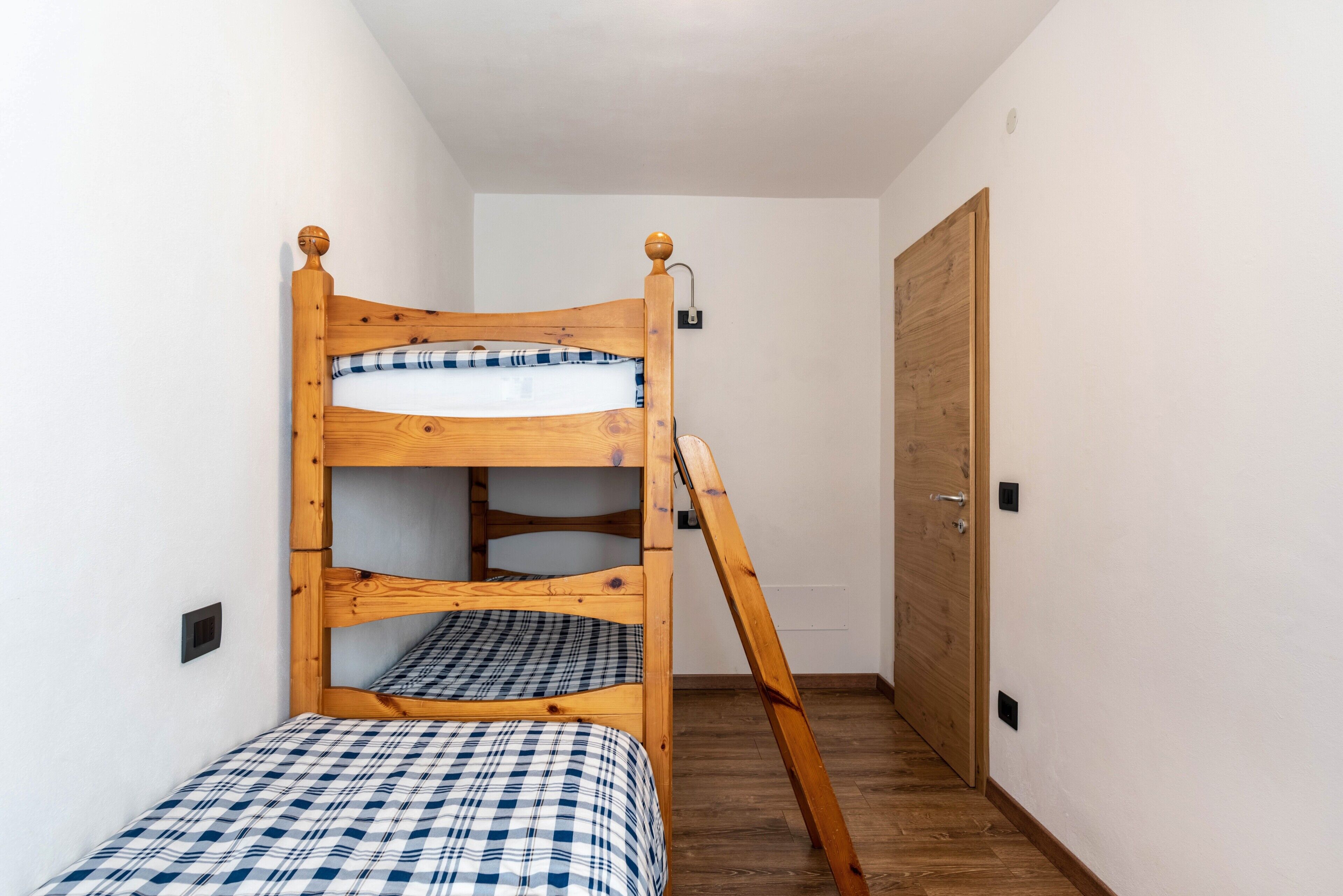 Ein Stockbett mit Holzrahmen und karierter Bettwäsche, eine Holztür und ein Lichtschalter an der Wand.