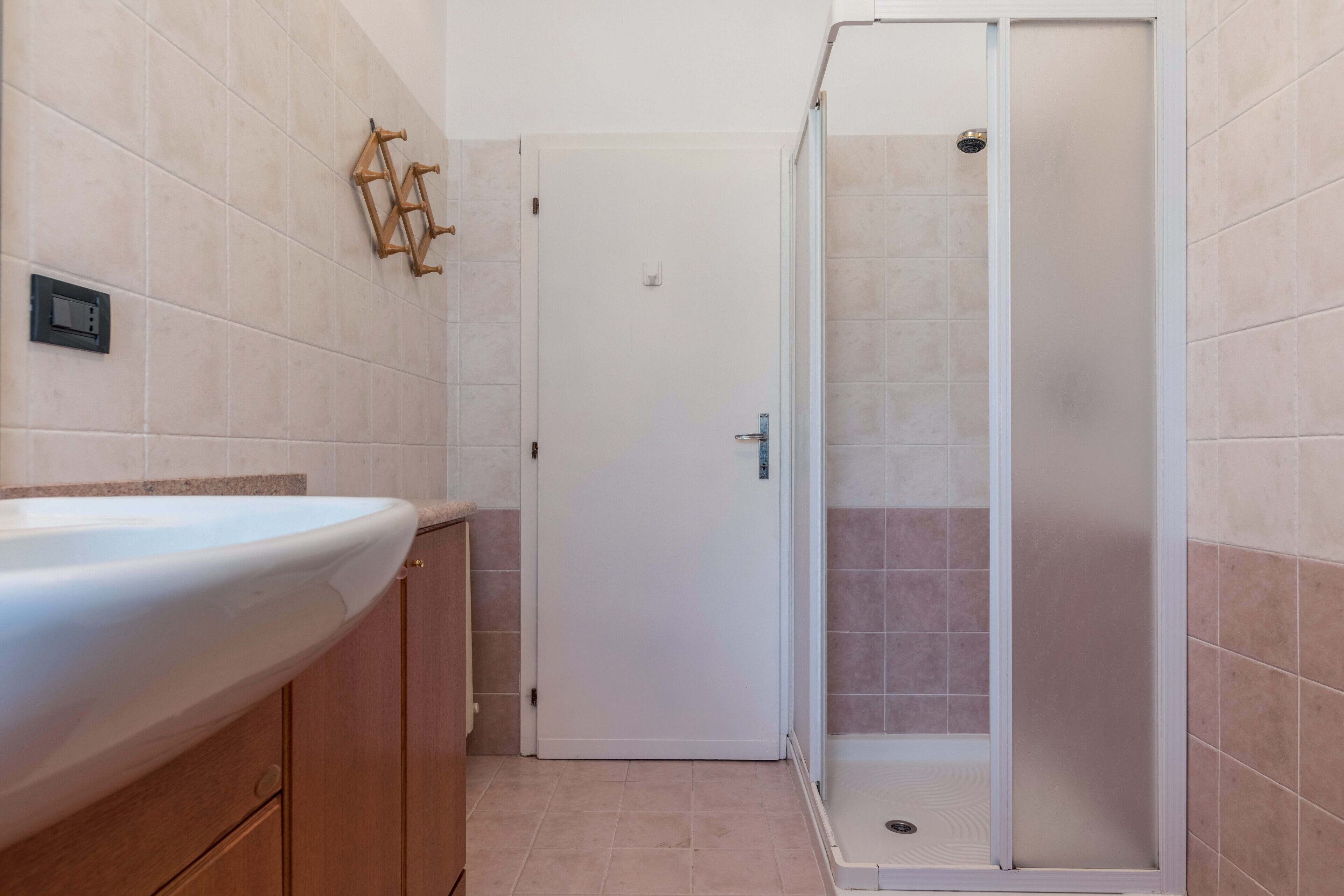 Un baño con un lavamanos blanco, armarios de madera, una puerta blanca y una zona de ducha con mampara de vidrio.