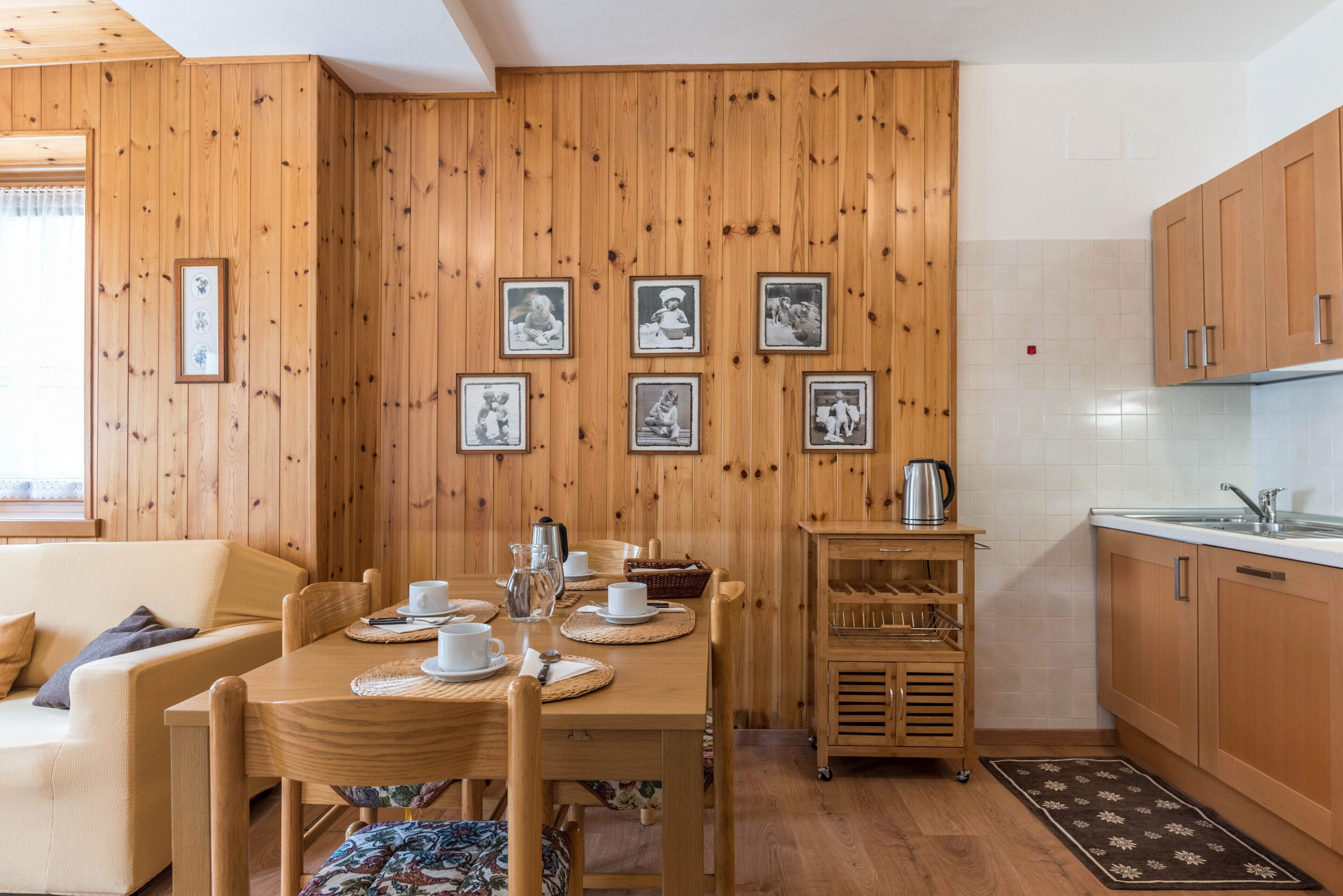 A wooden-paneled room with a dining table set for two, a kitchen area with wooden cabinets, and framed pictures on the wall.