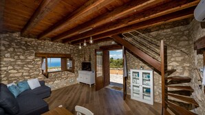 A rustic interior with stone walls, a wooden ceiling, a sofa, a TV, a staircase, and a view through a door.