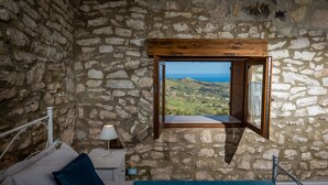 A stone-walled bedroom with a bed, a nightstand, a lamp, and a window with a view of the landscape.