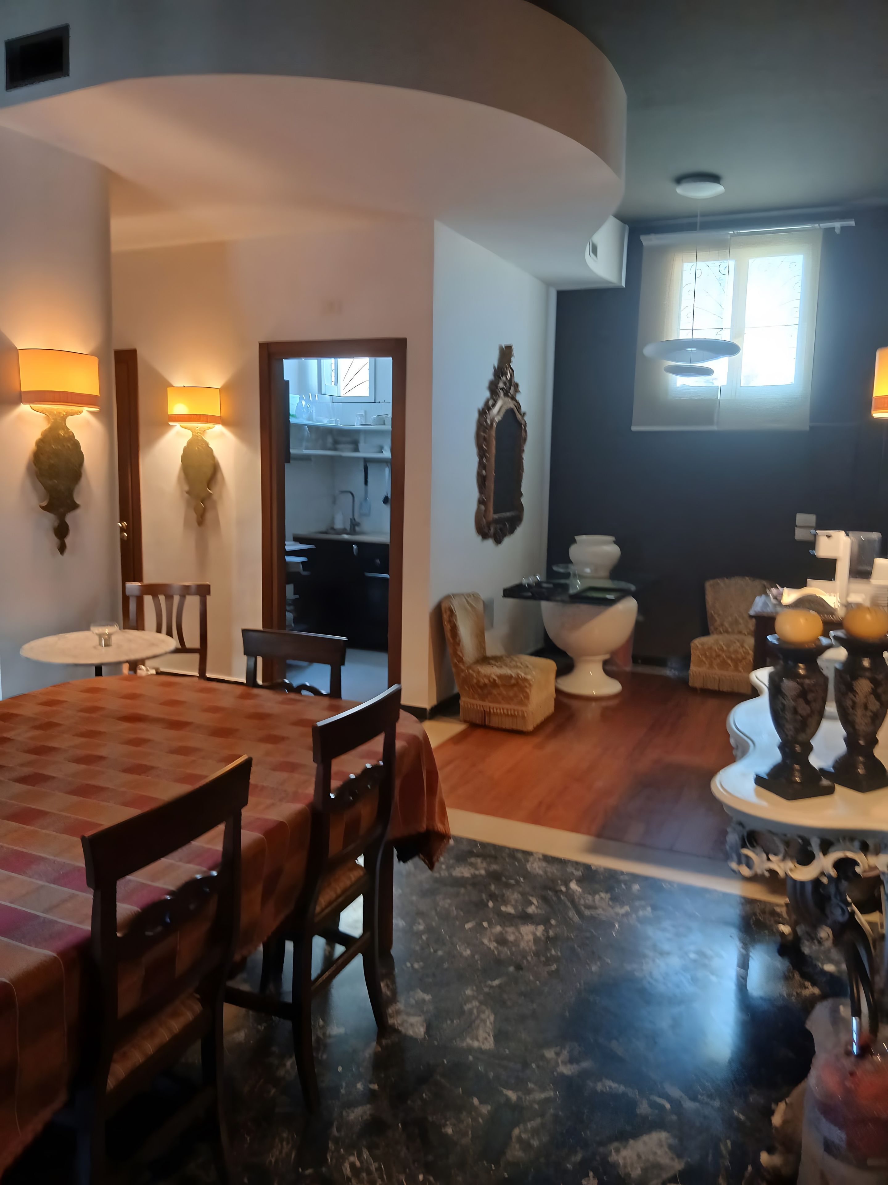 A dining area with a round table and chairs, a kitchen in the background, and a living area with a sofa and decorative items.