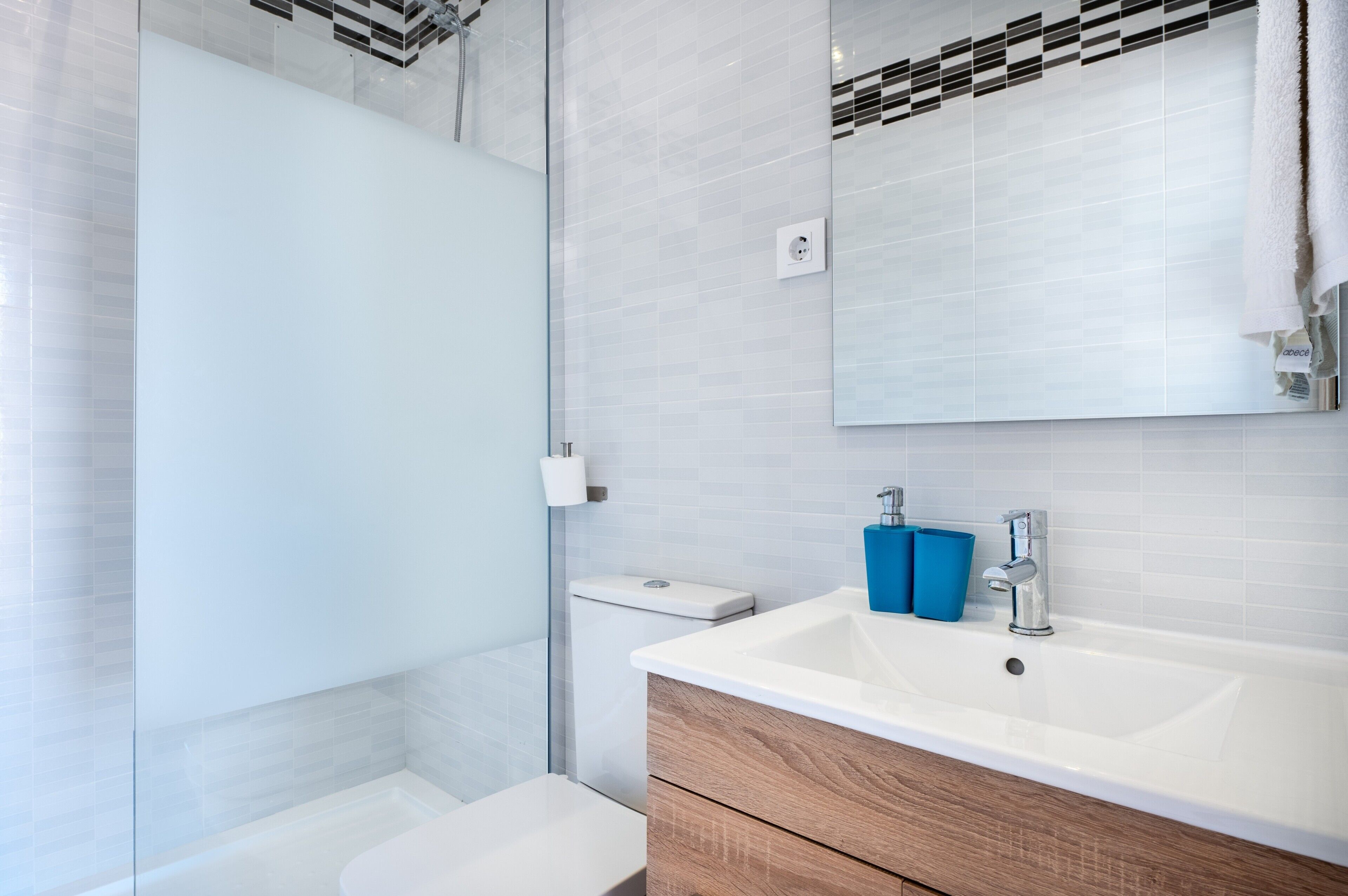 A modern bathroom with a glass shower enclosure, a white sink with a wooden vanity, and a mirror with a black and white tile backsplash.