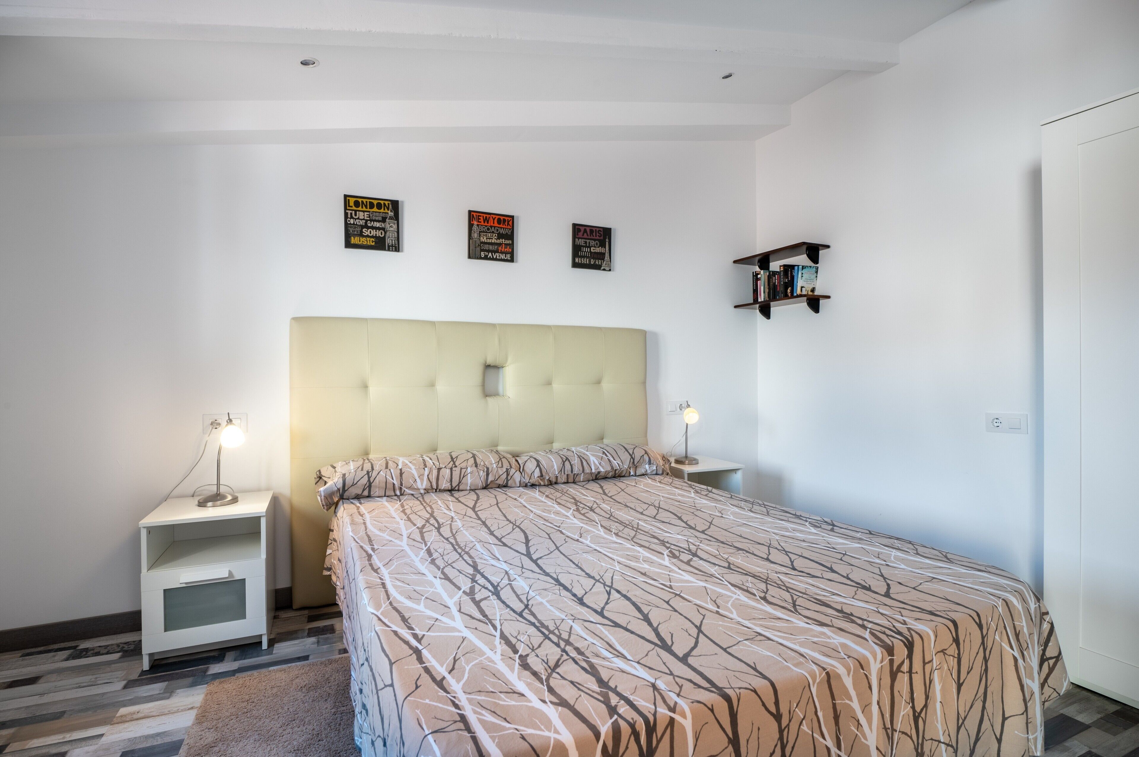 Une chambre à coucher avec un grand lit, deux tables de chevet, une étagère fixée au mur et trois affiches encadrées accrochées au mur.