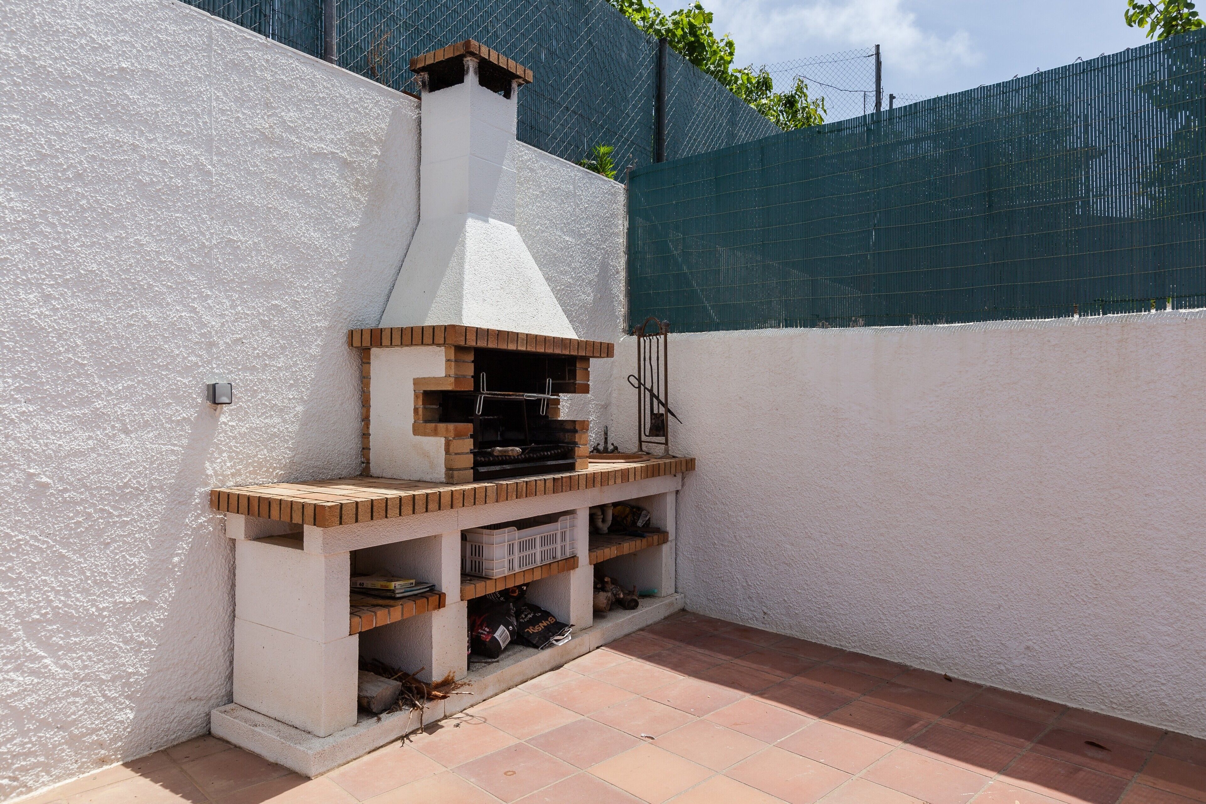 Un barbecue extérieur en briques, doté d’une cheminée, entouré d’un mur blanc et d’une clôture verte.