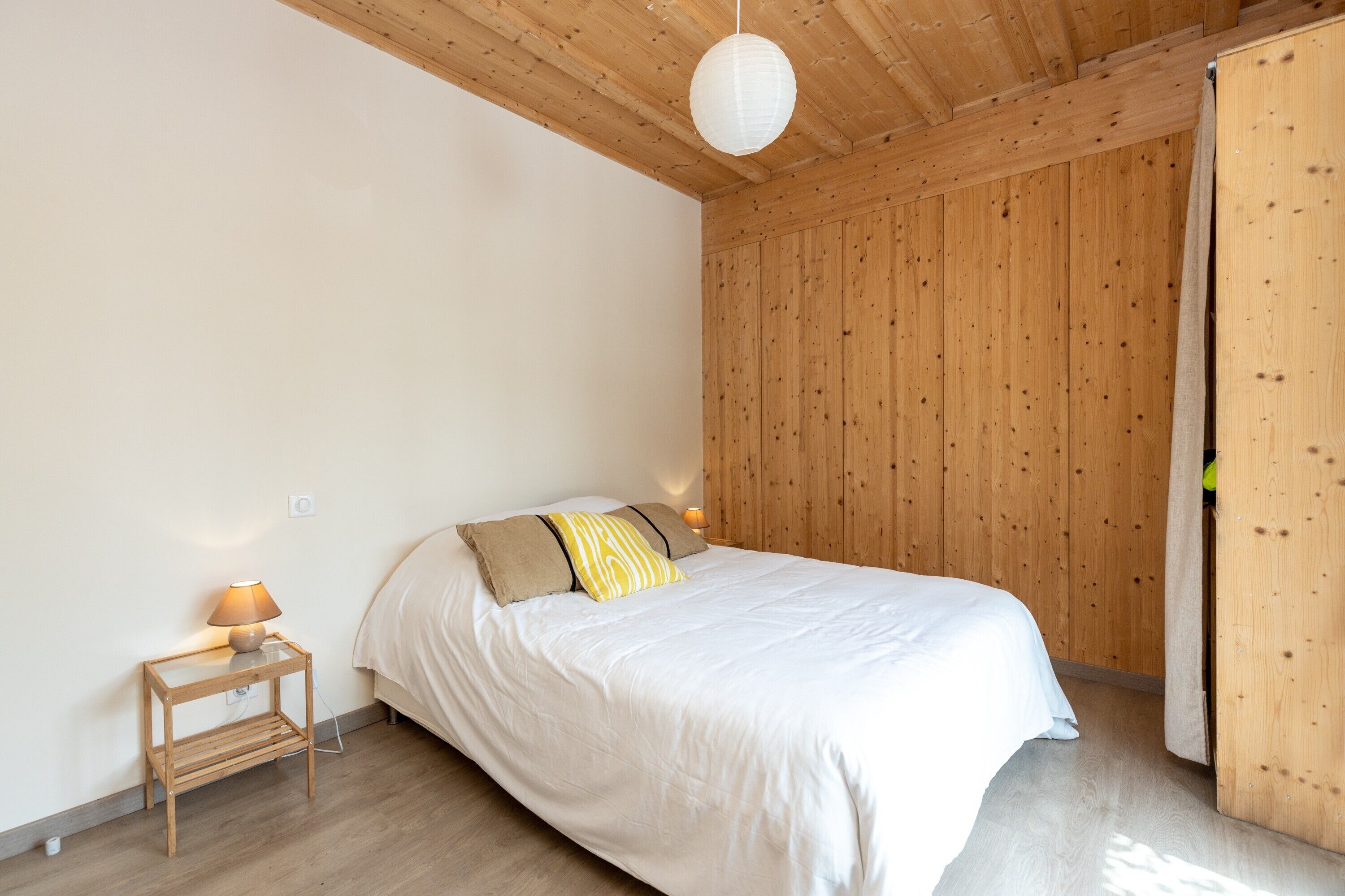 Een slaapkamer met een houten plafond, een bed met witte beddengoed, een nachtkastje met een lamp en een houten garderobe.