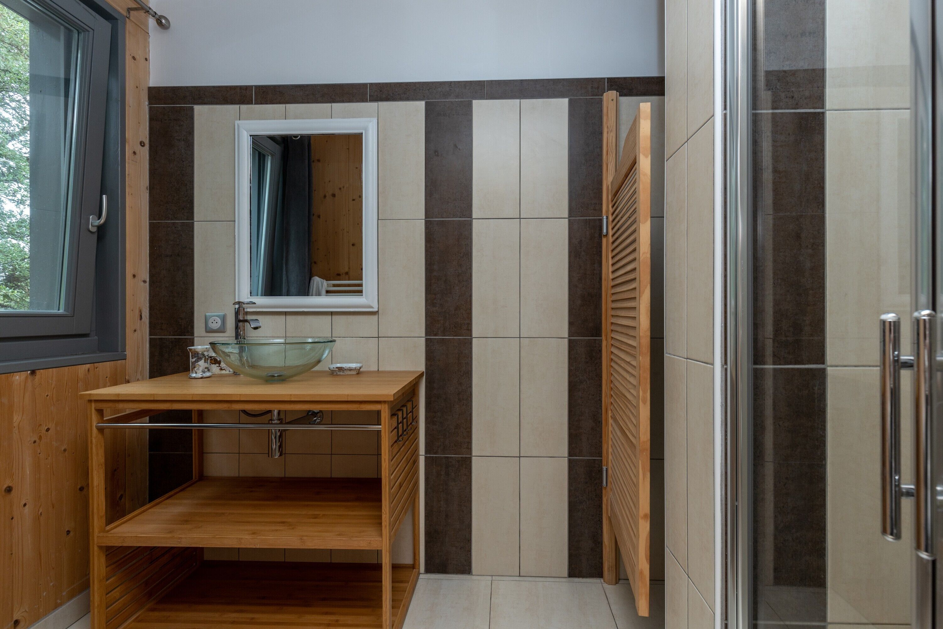 A bathroom with a wooden vanity, a glass bowl sink, a mirror, and a shower area.