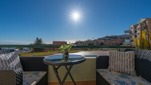 A sunlit outdoor seating area with a round table, cushioned chairs, and a view of a coastal town.