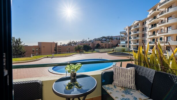A balcony with a view of a swimming pool, outdoor seating, and a building complex in the background.
