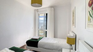 A bedroom with a bed, a bedside table, a lamp, and a window with a view of the city. - Lido Life Apartment by LovelyStay (Funchal)