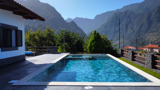 A rectangular swimming pool with clear blue water, surrounded by a stone deck. In the background, there are mountains and a building with a red roof.
