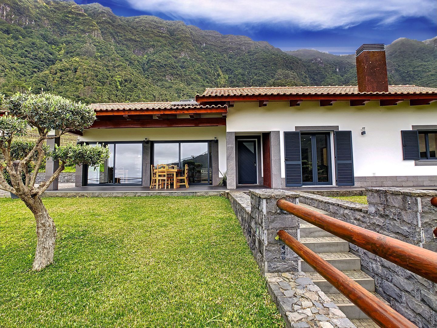 A modern house with a stone staircase leading to a wooden deck, surrounded by lush greenery and mountains.