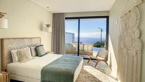 A bedroom with a large bed, a view of the ocean, and a macrame wall hanging. - Noah - Private Sunset Hideaway (Arco Da Calheta)