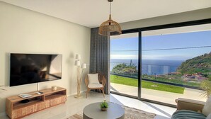 A modern living room with a large TV, a round coffee table, and a view of the ocean. - Noah - Private Sunset Hideaway (Arco Da Calheta)