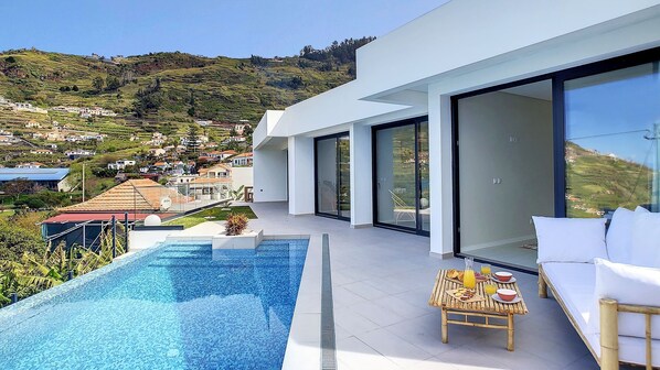 Een modern huis met een zwembad, een eethoek met een tafel en stoelen, en een grote glazen deur die leidt naar een balkon met uitzicht op een heuvelstad.