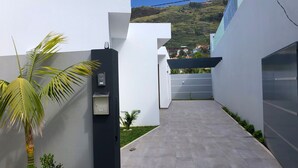A narrow, paved pathway leading to a modern building with a flat roof and a view of a hilly landscape in the background. - Noah - Private Sunset Hideaway (Arco Da Calheta)