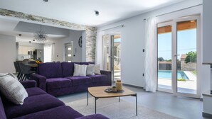 Una sala moderna con un sofá morado, una mesa de centro de madera y vista a una piscina a través de grandes puertas corredizas de vidrio.