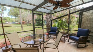 A covered patio area with a glass table and chairs, a ceiling fan, and a view of the surrounding garden.