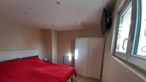 Un dormitorio con una colcha roja, un armario, un televisor montado en la pared y una ventana con persianas.