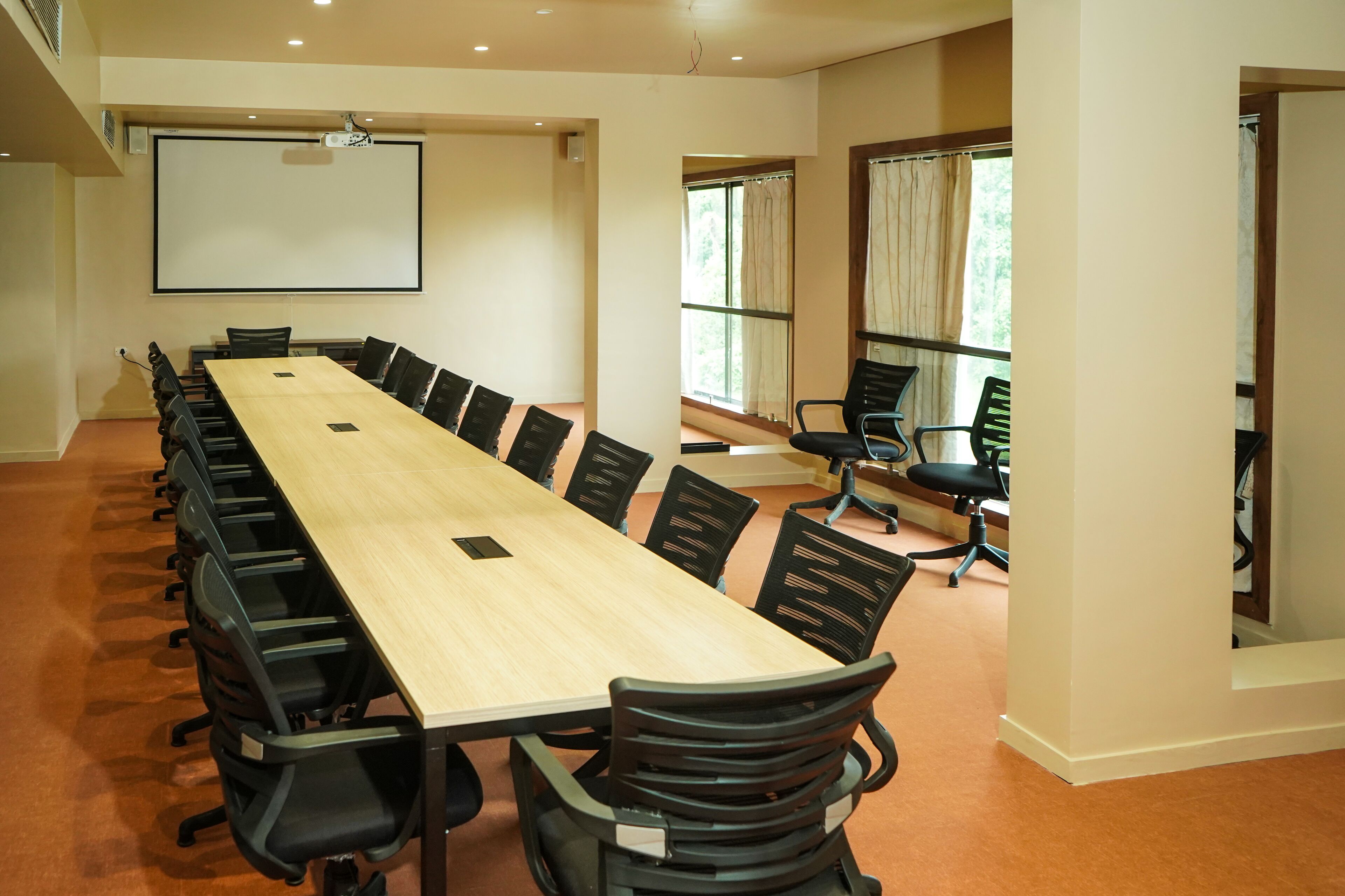 A conference room with a long table, black chairs, a projector screen, and windows with curtains.