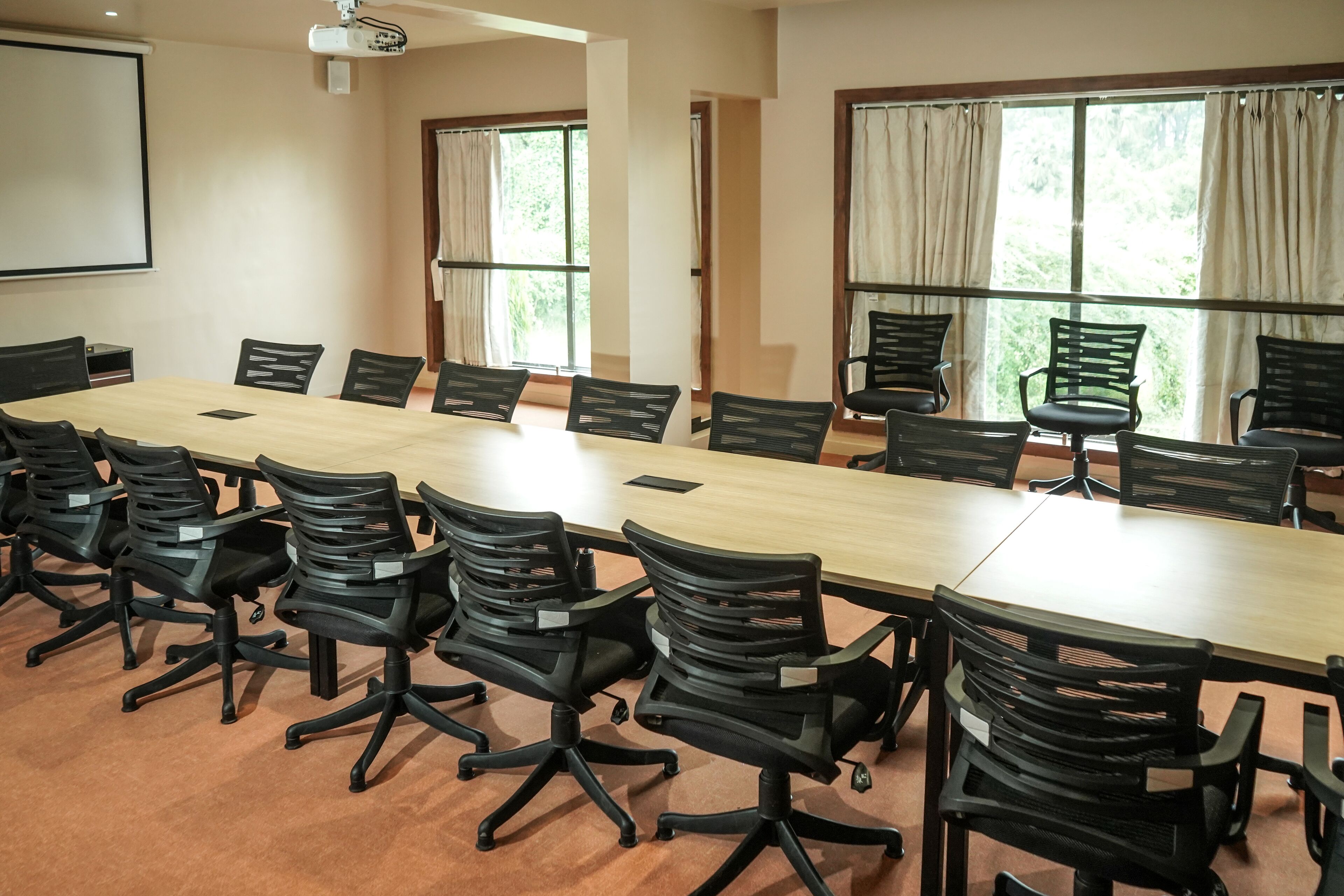 A conference room with a long table, black chairs, a projector screen, and large windows with curtains.