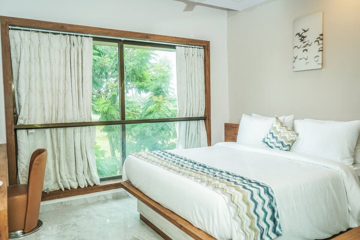 Ein Schlafzimmer mit großem Fenster, einem Bett mit weißer Bettwäsche und Blick auf Grün außerhalb.