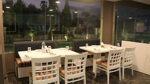 Un restaurante con mesas y sillas blancas, suelo de mármol y vistas a árboles y un edificio a través de amplias ventanas.